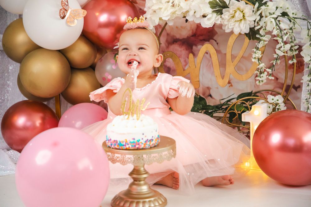 Baby girl in pink tutu smashing her first birthday cake surrounded by pink and gold balloons and floral decorations.