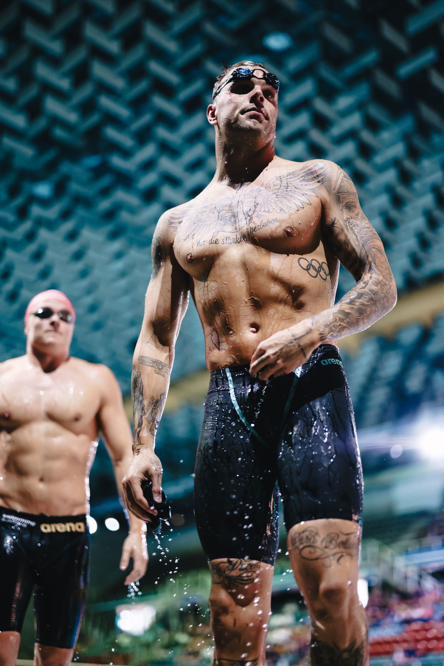 Tattooed swimmer in black swimsuit emerging from pool after race.