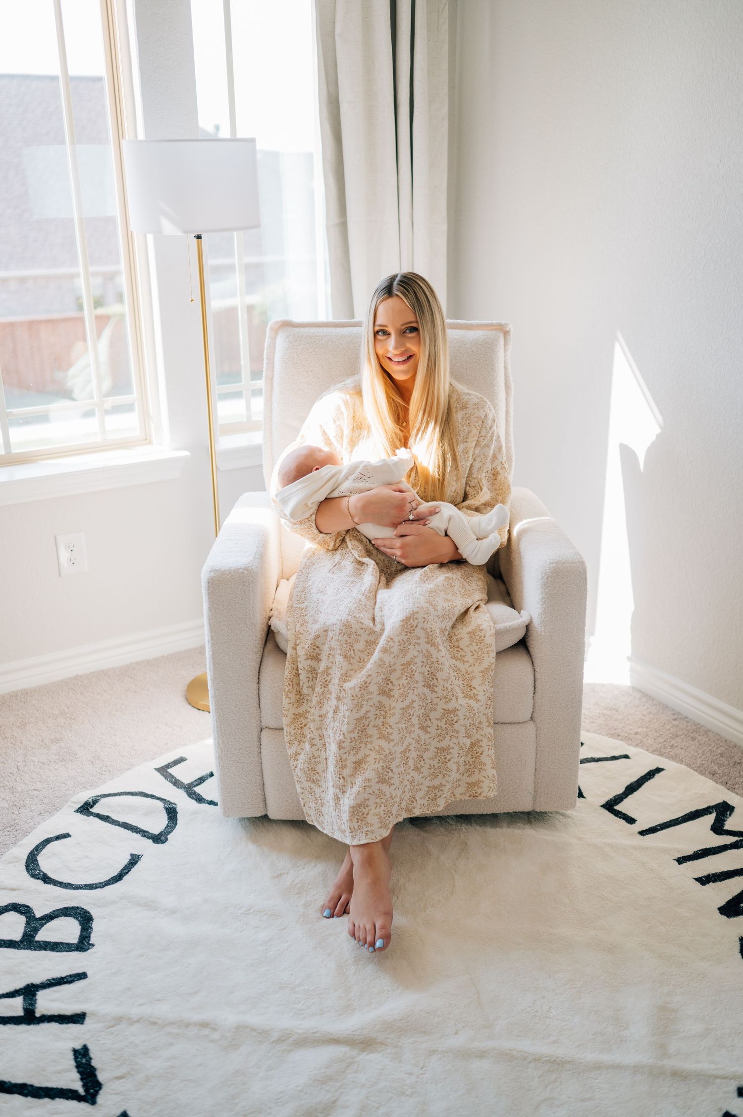 Mother in cream dress sitting in white chair with newborn on alphabet rug.