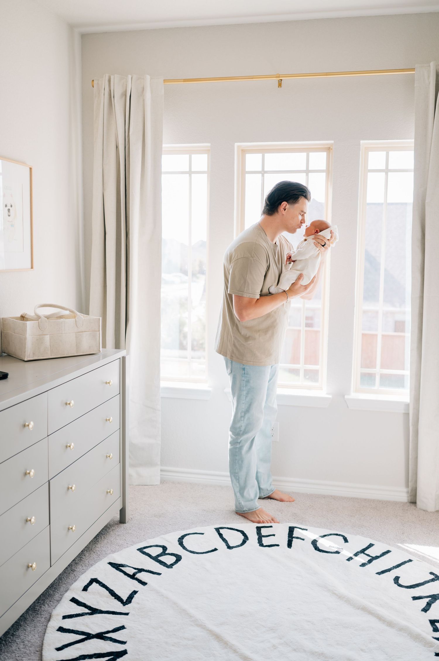Parent holds newborn baby while standing by window in modern nursery with alphabet rug.