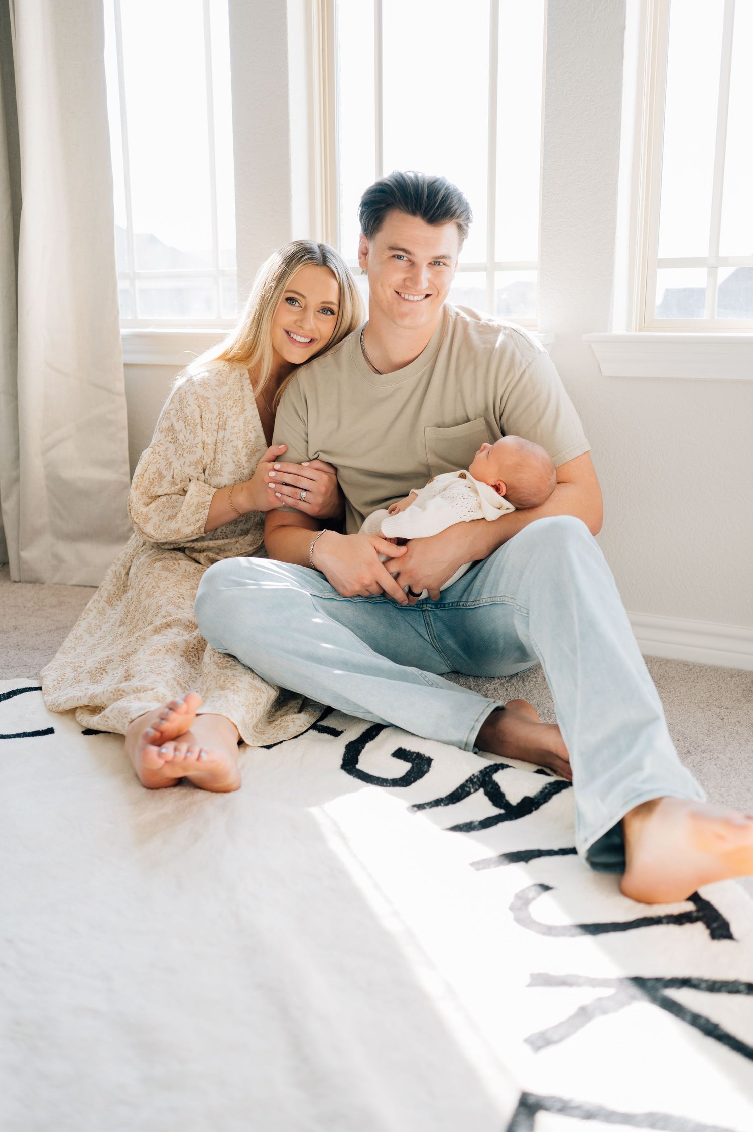 Happy new parents sit with their newborn baby on alphabet play mat in sunny nursery.