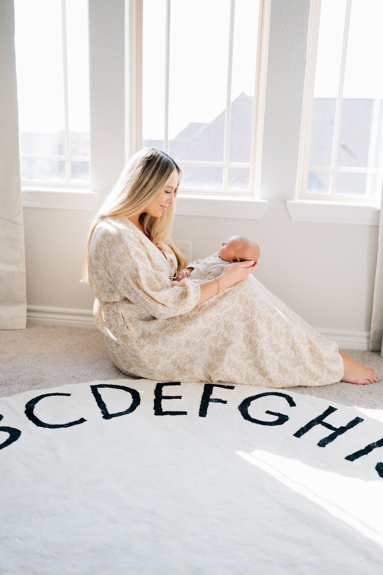Parent sits on alphabet rug holding newborn baby in cream floral dress with natural window light.