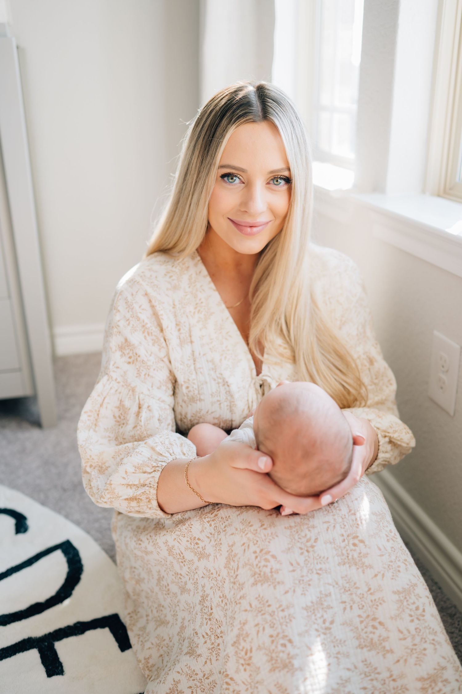 Mom holding and gazing at sleeping newborn on alphabet rug in cream floral dress.