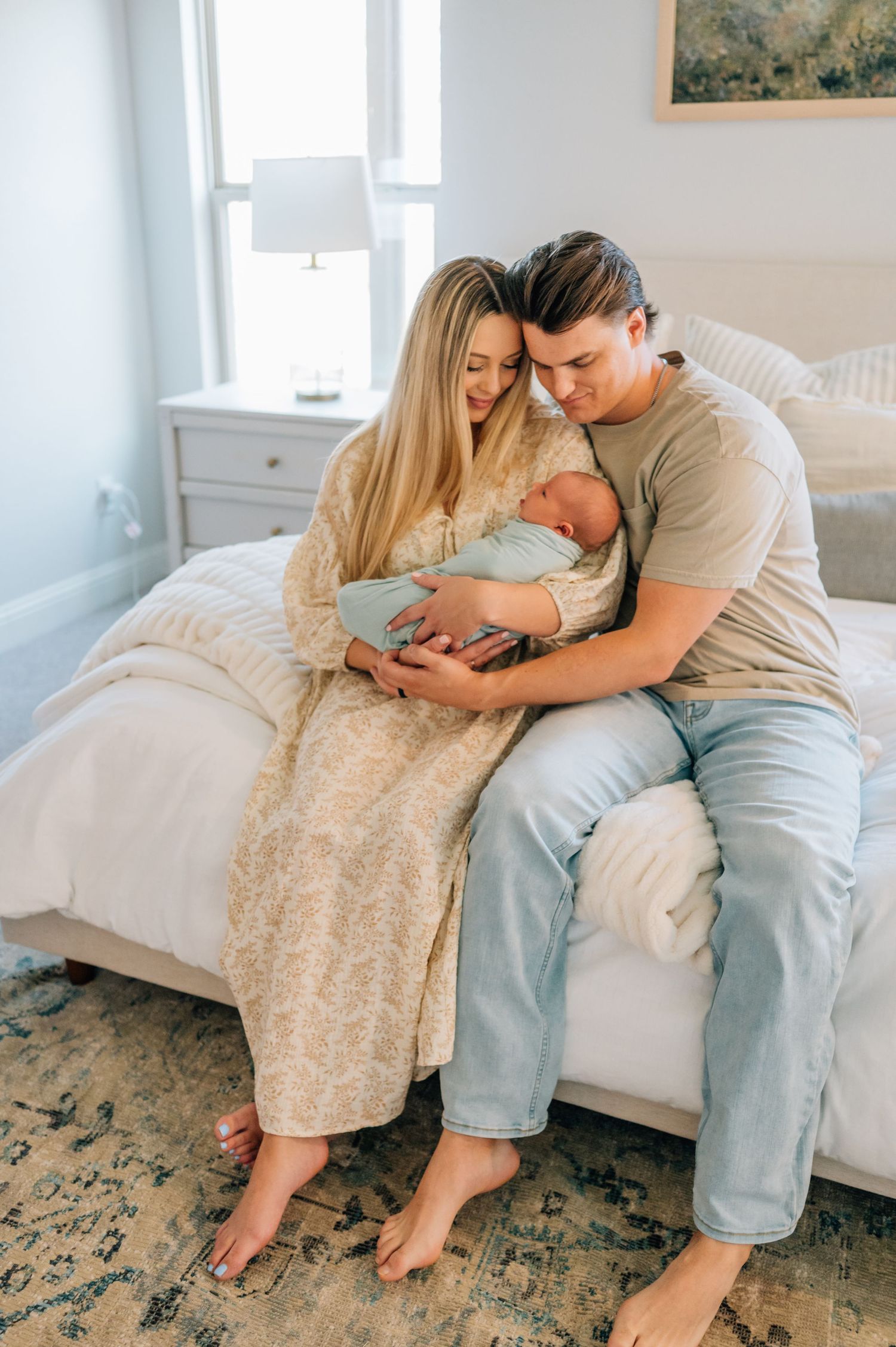 Parents tenderly holding their newborn baby while sitting on a white bed in a bright, minimalist bedroom.