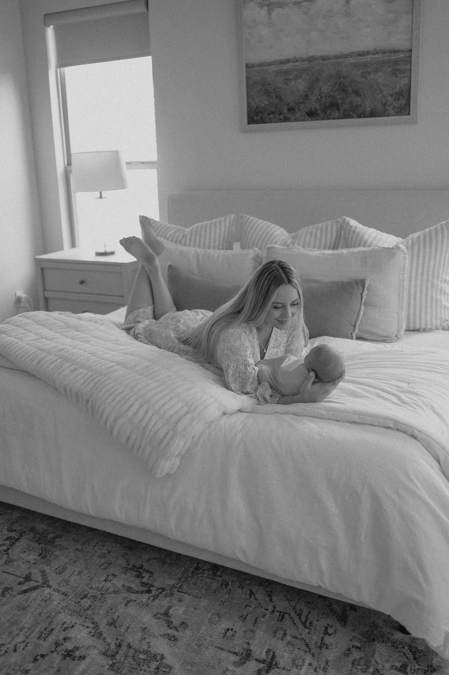 Mother lying on bed holding newborn in black and white photo with soft window light.