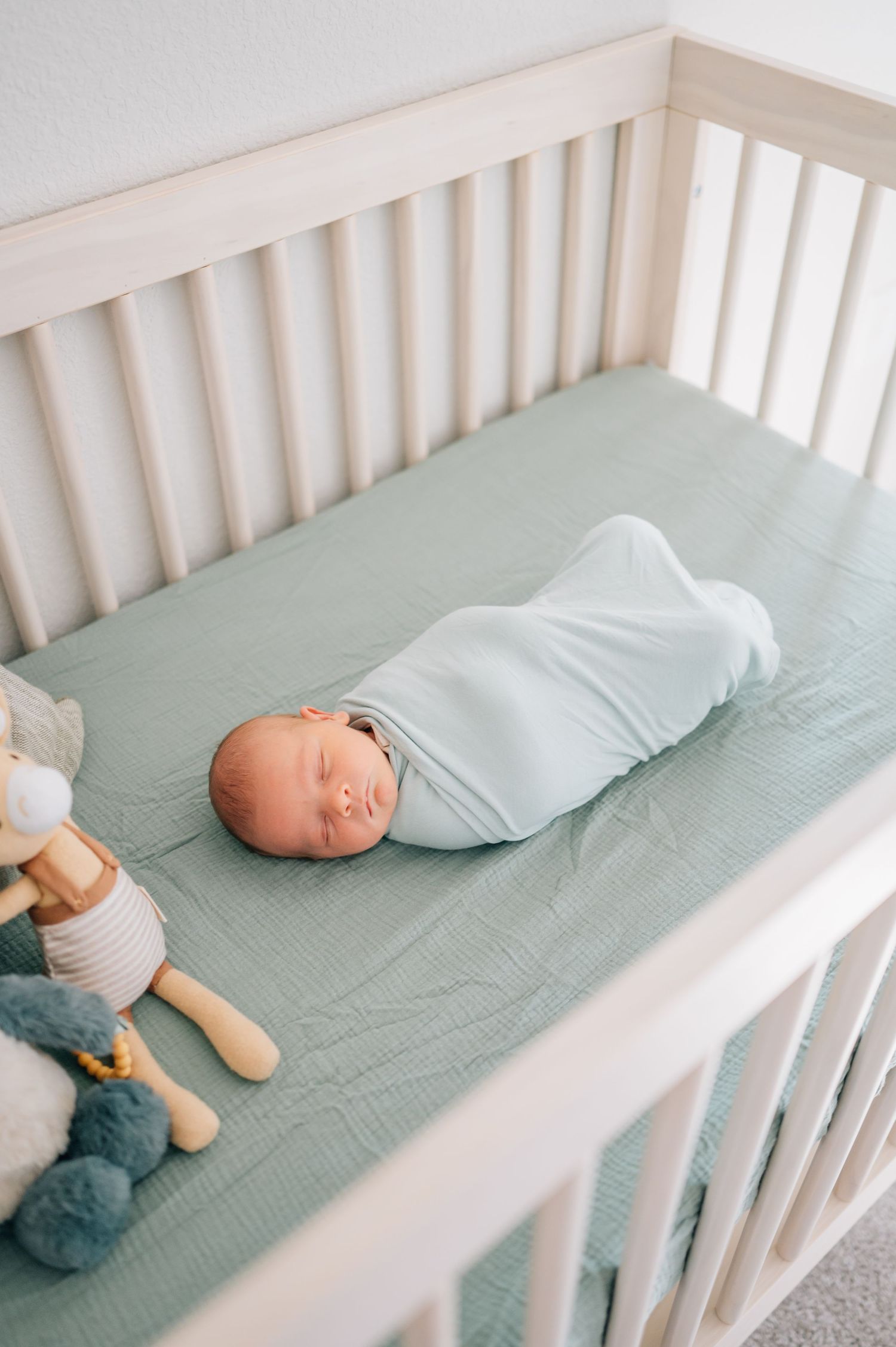 Sleeping swaddled newborn in white crib with stuffed animals and mint green sheet.