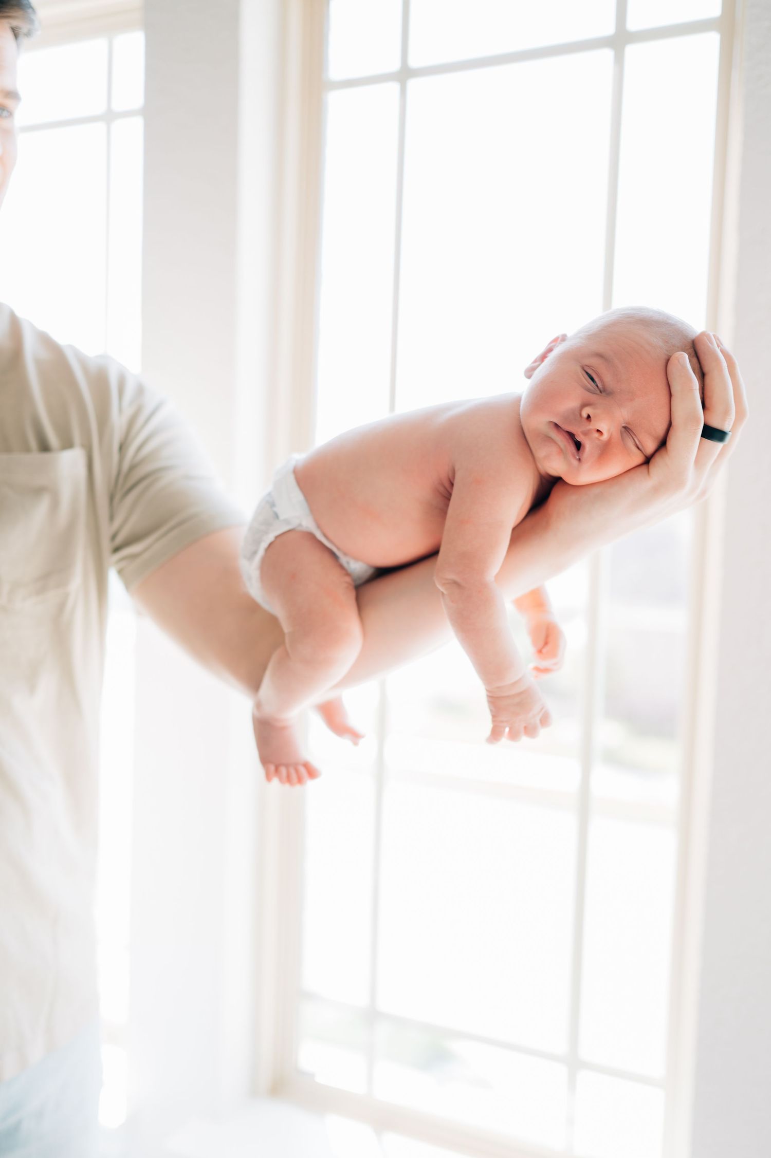 Parent holding sleeping newborn baby against bright window in natural light.