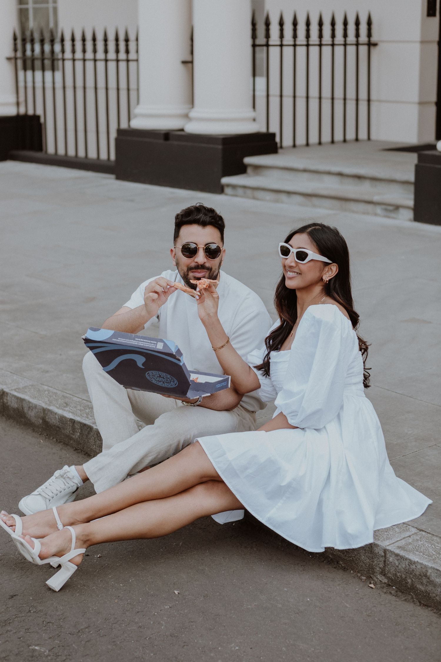 Stylish couple in white outfits and sunglasses sitting on urban steps sharing playful moment.