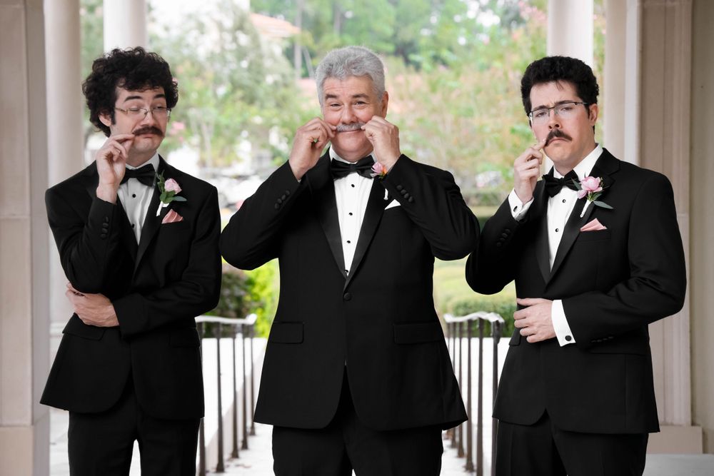 Three men in black tuxedos playfully twirl their mustaches while posing together.