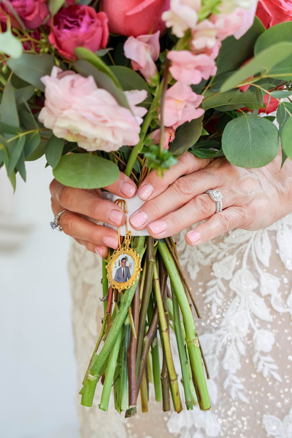 Bridal bouquet of pink roses with memorial locket attached being held by lace-covered hands.