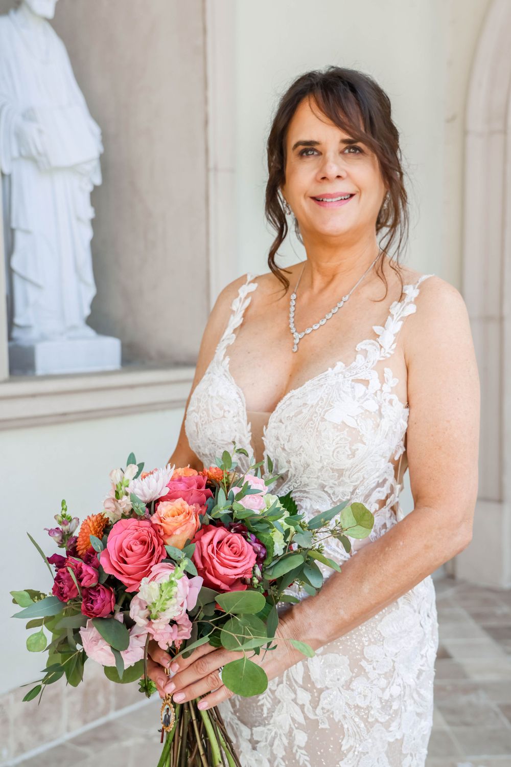 A bride in a detailed lace wedding dress smiles while holding a vibrant pink and coral rose bouquet against a classic backdrop.