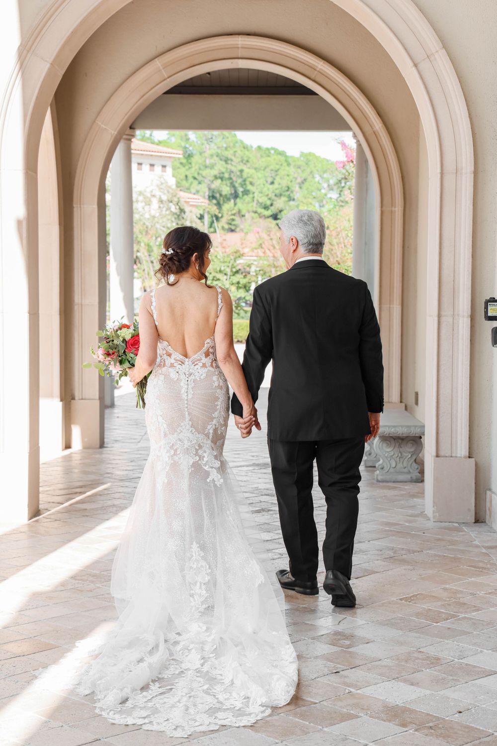Back view of a bride in a lace gown walking through an arched hallway holding hands with a companion.