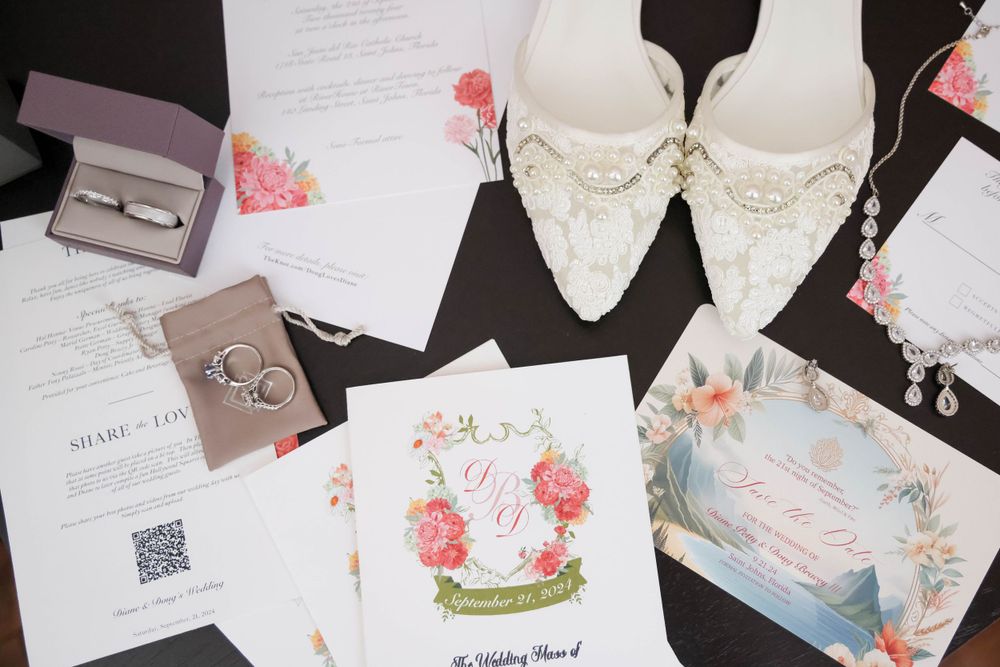 Wedding invitations, rings and elegant white bridal shoes with pearl details arranged on dark surface.