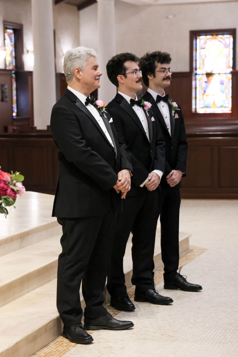 Three men in formal tuxedos stand on church steps during wedding ceremony.