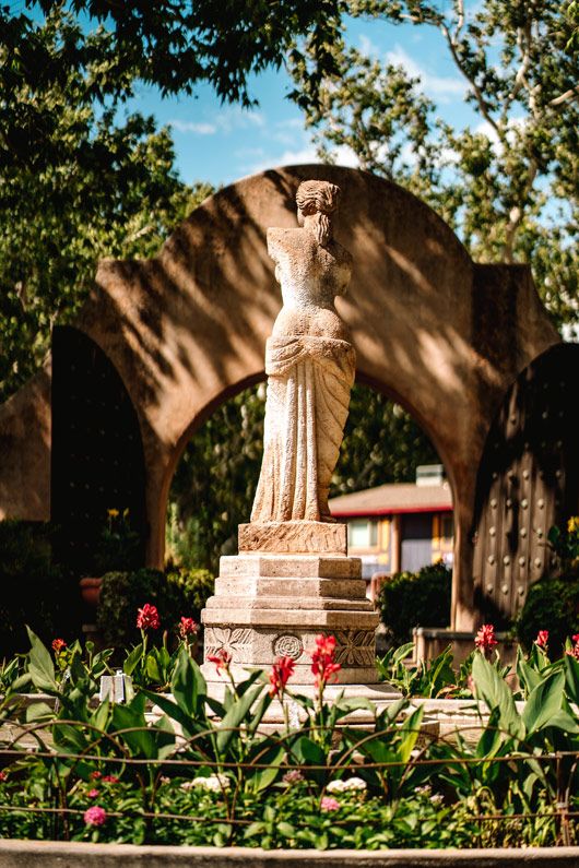 Classical Sedona statue surrounded by red flowers and arched architecture in a garden setting.