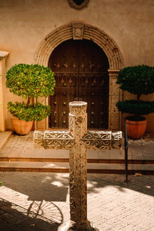 Ornate Sedona cross standing in front of a historic wooden door with potted trees on either side.