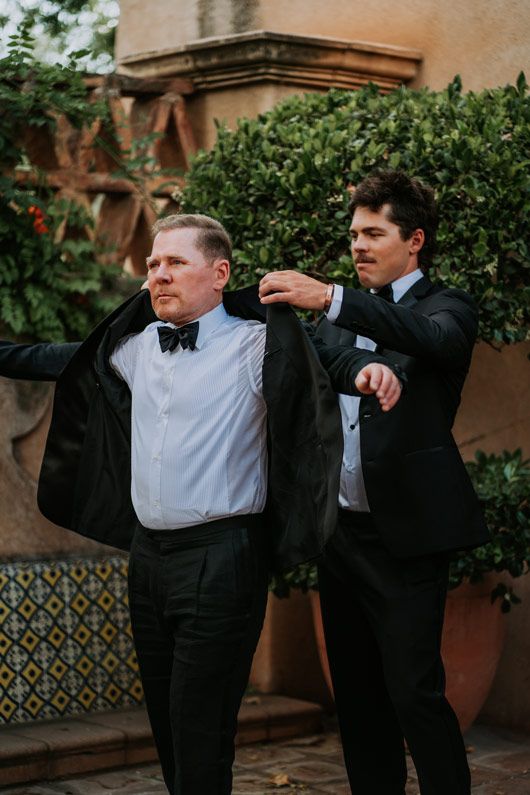 Two people in formal attire help each other adjust their suits outdoors.