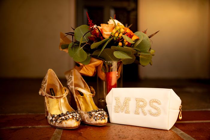 Wedding accessories including ornate gold heels and a 'MRS' sign displayed next to flowers in warm lighting.