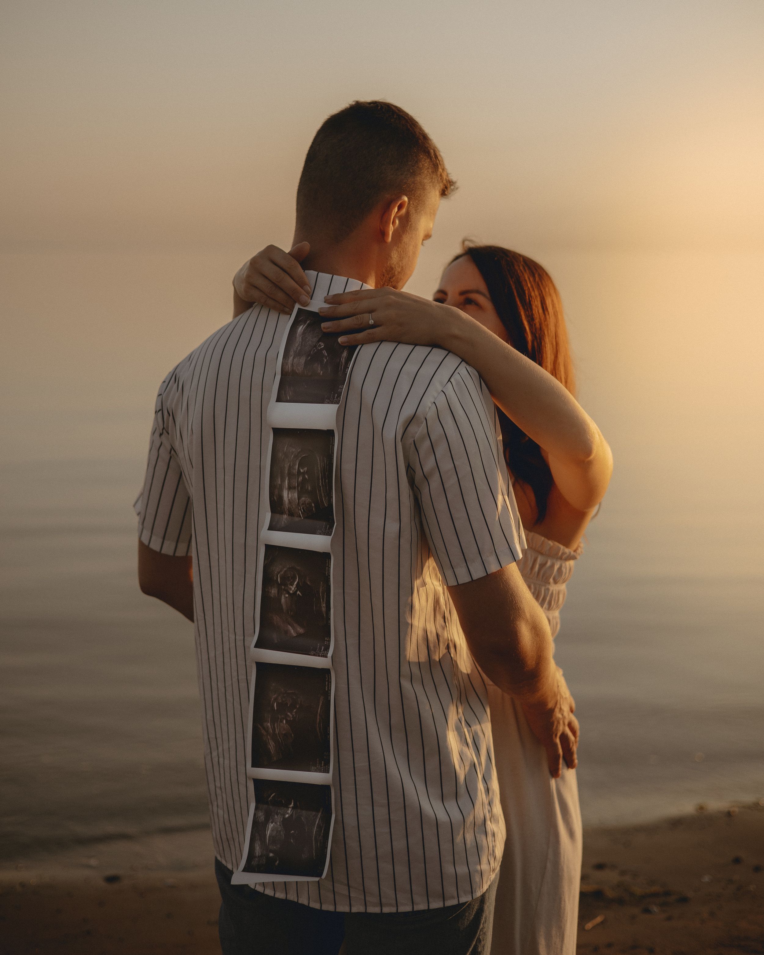 A couple shares an intimate moment while holding ultrasound photos at sunset by the water.