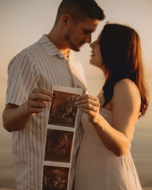 A couple shares a tender moment while holding sonogram photos during golden hour.