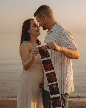 A couple shares an intimate moment while holding ultrasound photos by the water at sunset.