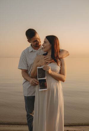 A couple shares a loving moment while holding sonogram photos at the beach during sunset.