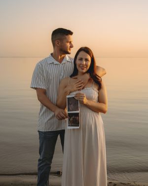 A couple poses with ultrasound photos during a sunset maternity photoshoot by the water.