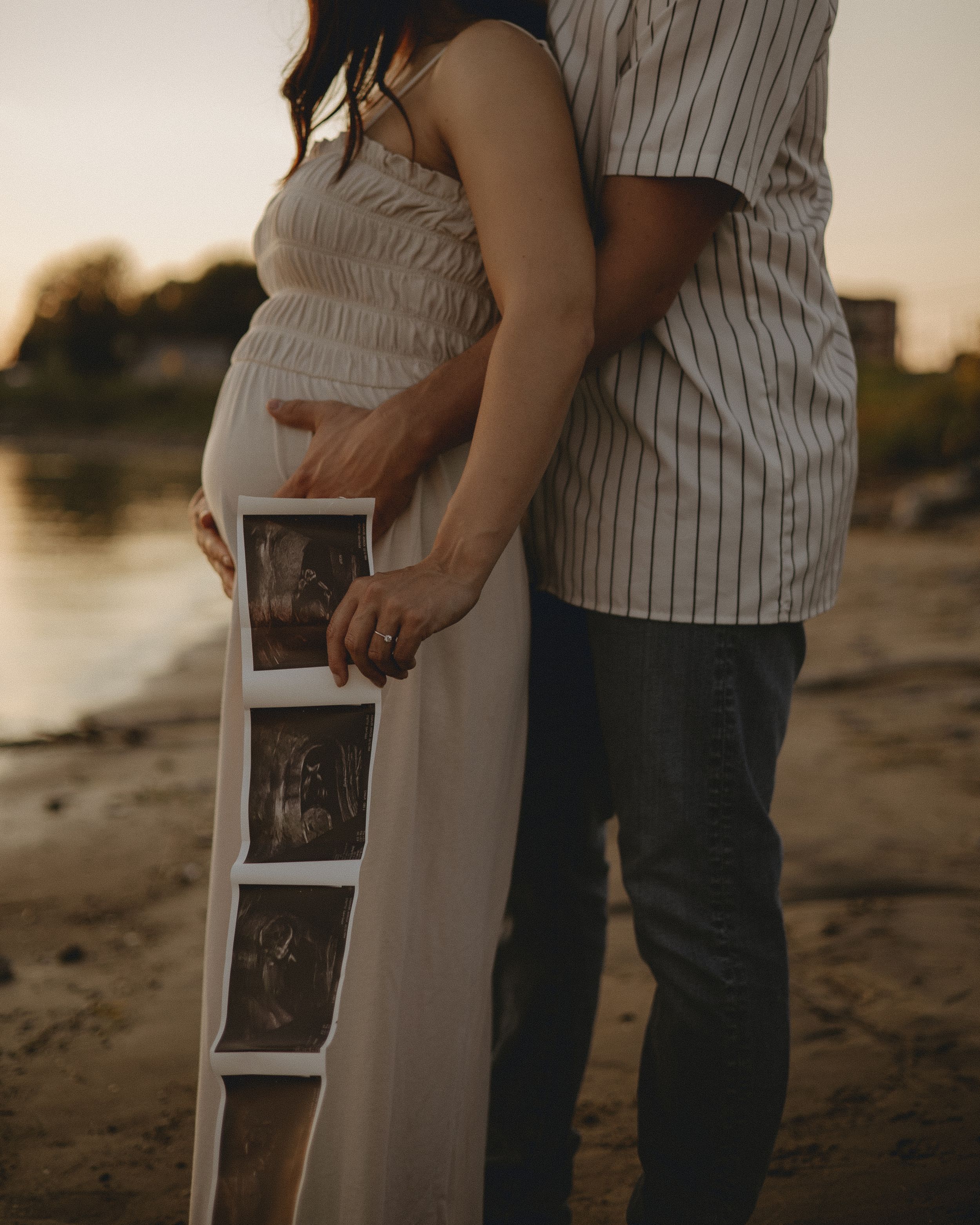 A couple on a beach holds ultrasound photos while embracing during sunset.