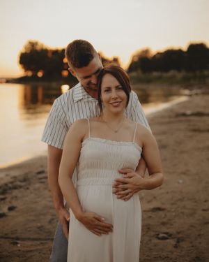 Couple embraces on beach at sunset during maternity photoshoot in white dress and striped shirt.