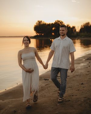 Expectant couple walks hand in hand along shoreline during golden hour sunset.