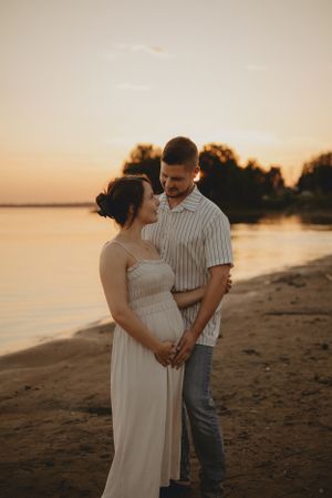 Intimate beach maternity photo with couple in white dress and striped shirt at dusk.