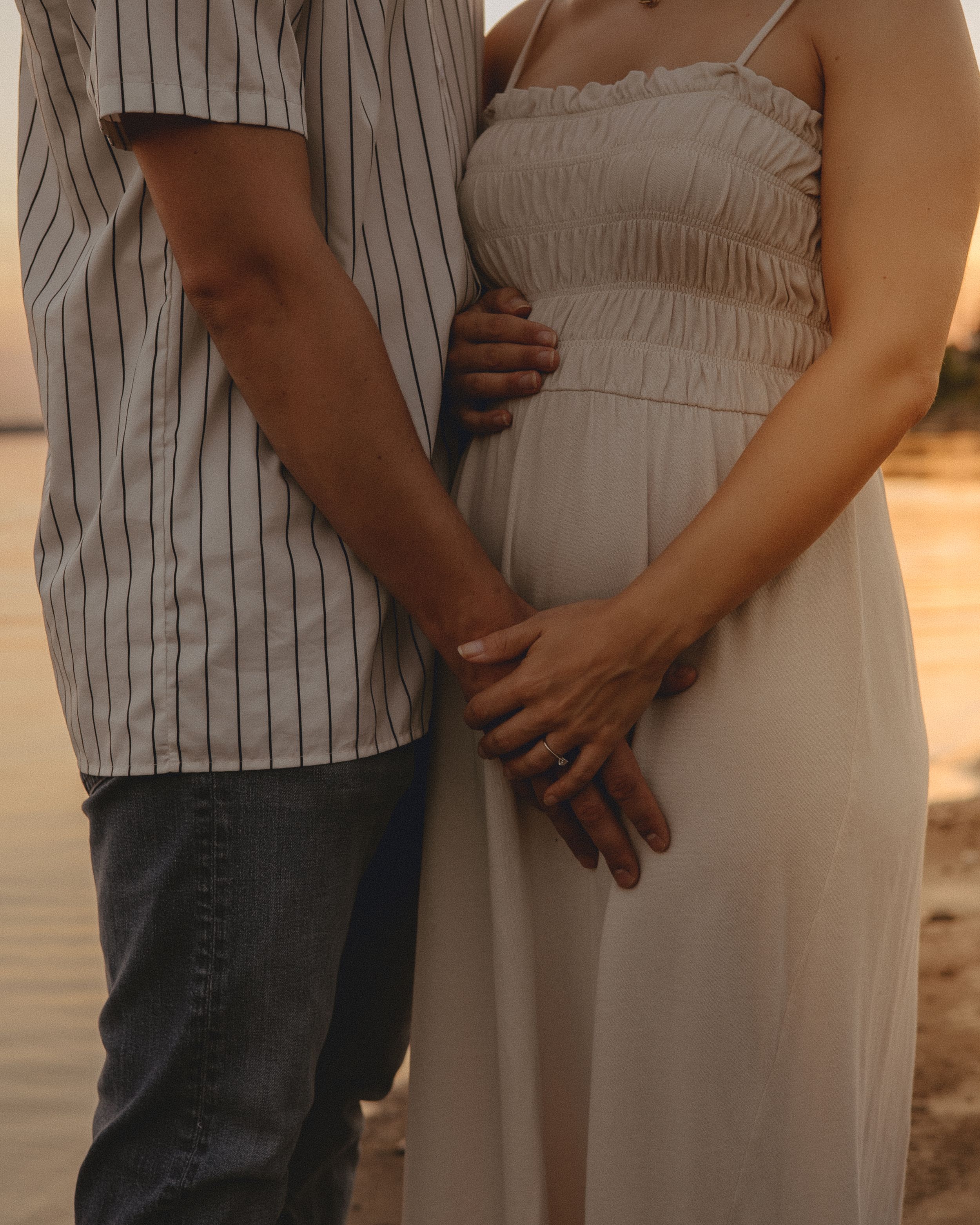 Close-up of couple's hands on maternity bump in white dress at golden hour.