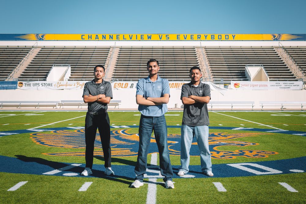 Three people stand together on a football field with stadium seating visible in the background.