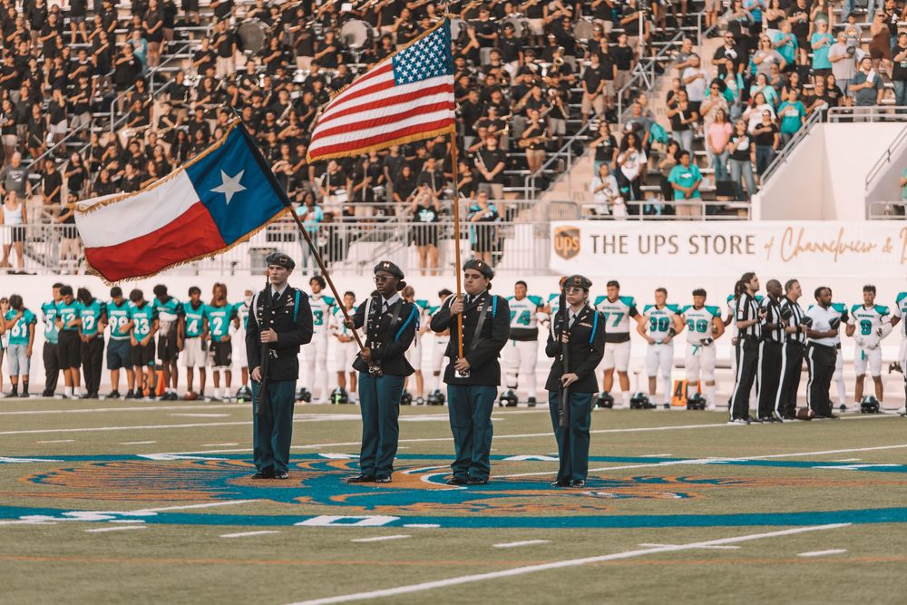Marching band members perform during a football game with American and Texas flags on display.