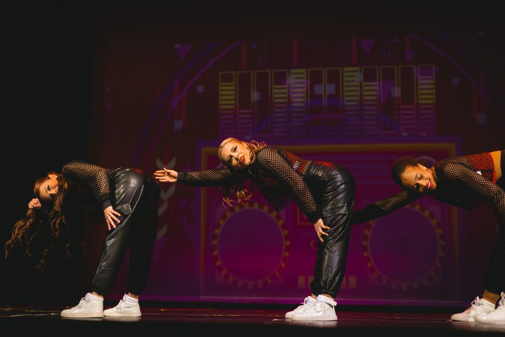 Three dancers in black outfits and white sneakers perform a synchronized lean on a colorful stage.