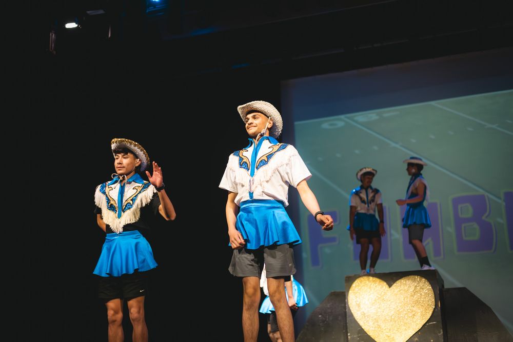 Children in blue and white costumes perform on stage with a heart decoration and colorful backdrop.