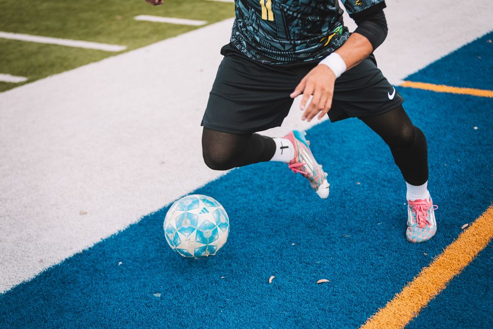 Soccer player in Nike gear kicking a ball on a blue turf field wearing pink cleats.
