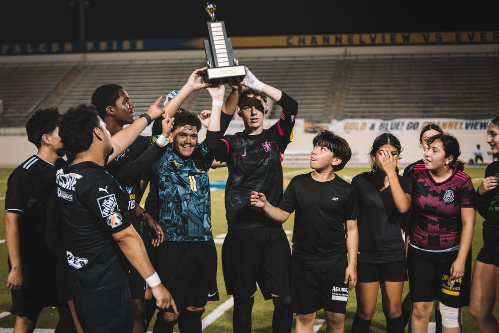 Soccer team celebrating championship victory at night, players raising trophy on field in black uniforms.