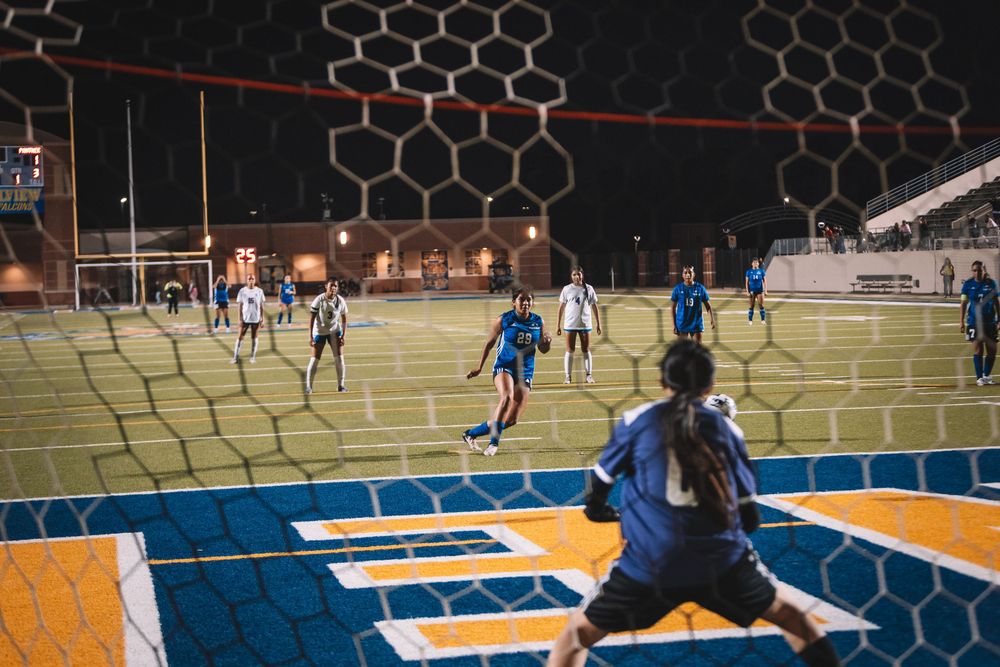 Indoor soccer players compete in a nighttime match viewed through a goal net with yellow and blue court markings.