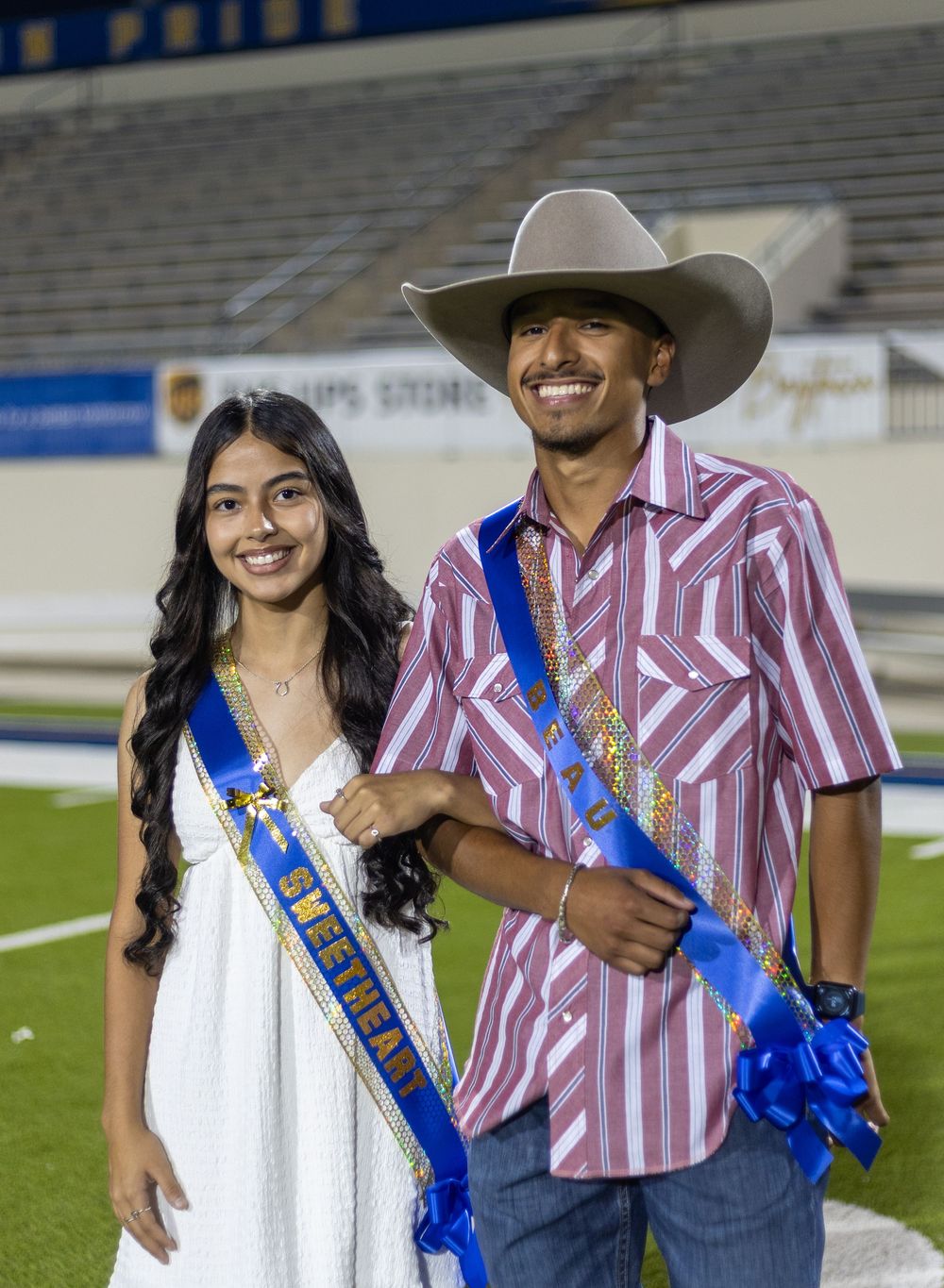 Homecoming couple poses on football field in western formal wear with sashes.