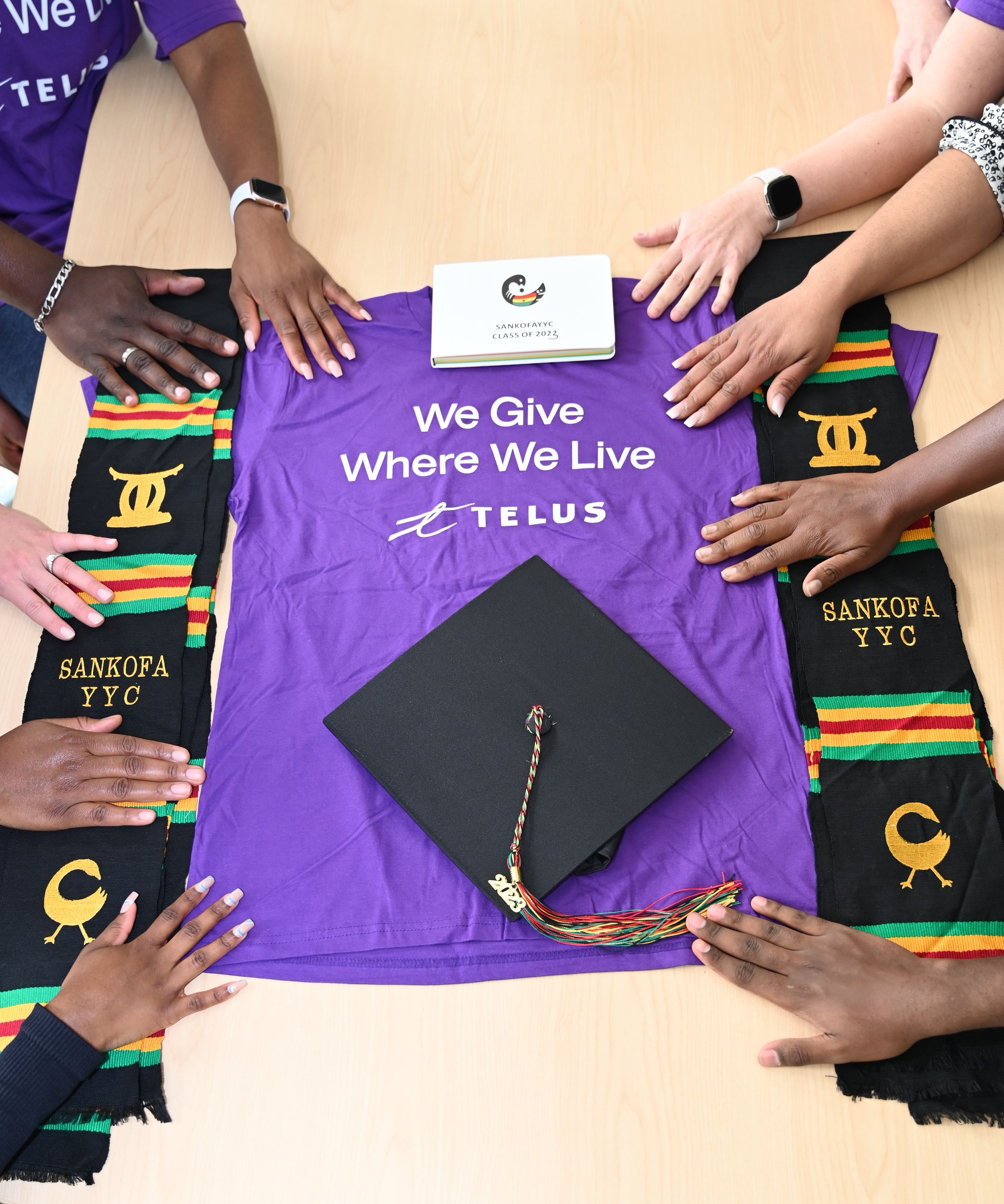 Corporate Events - Telus Day of Giving. Hands touching Kente scarfs surrounding a purple t-shirt that says We Give Where We Live Telus