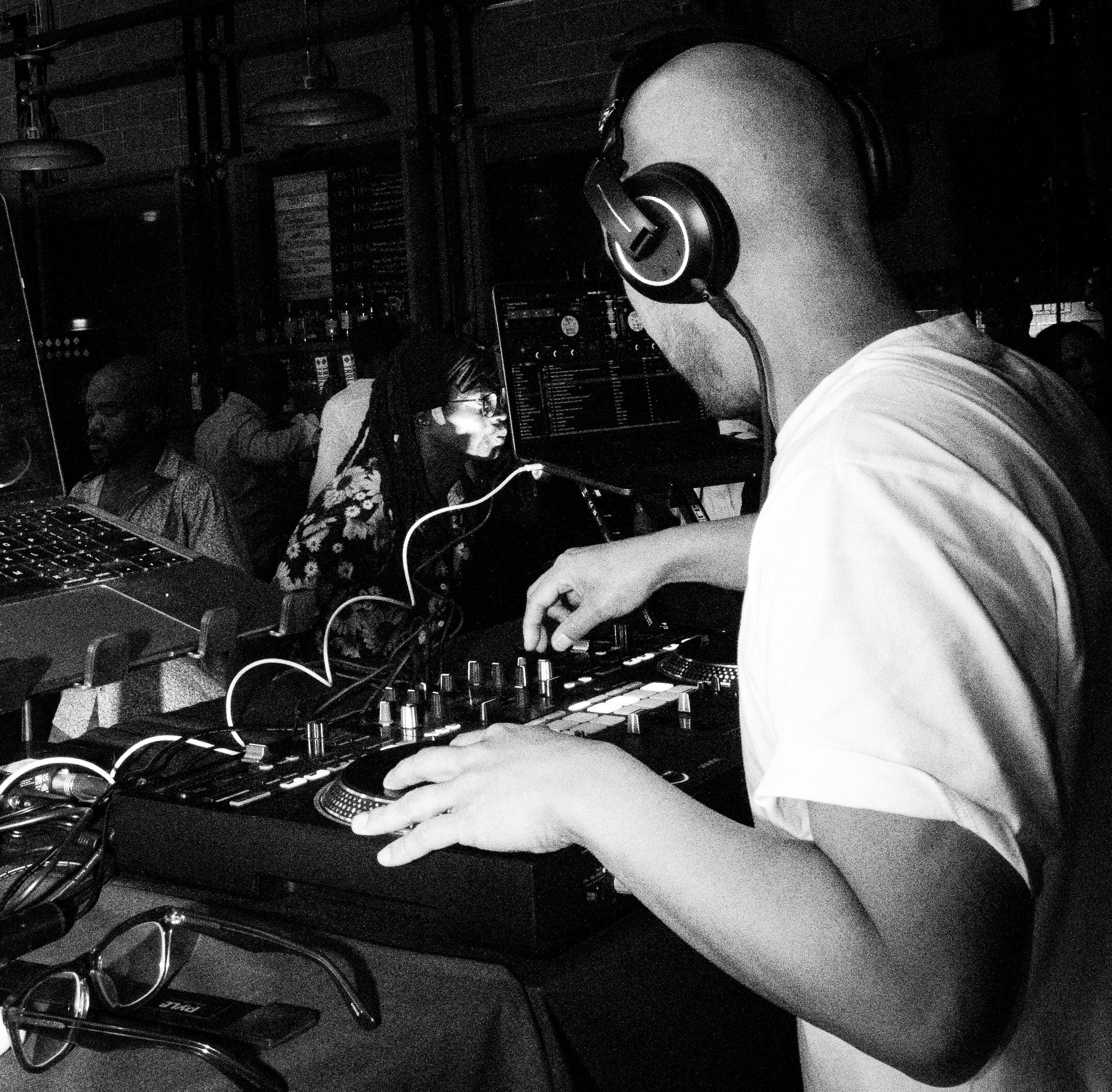 A black and white photo of a Filipino DJ on a slightly raised stage. He adjusts his controller as the crowd dances to the beat.