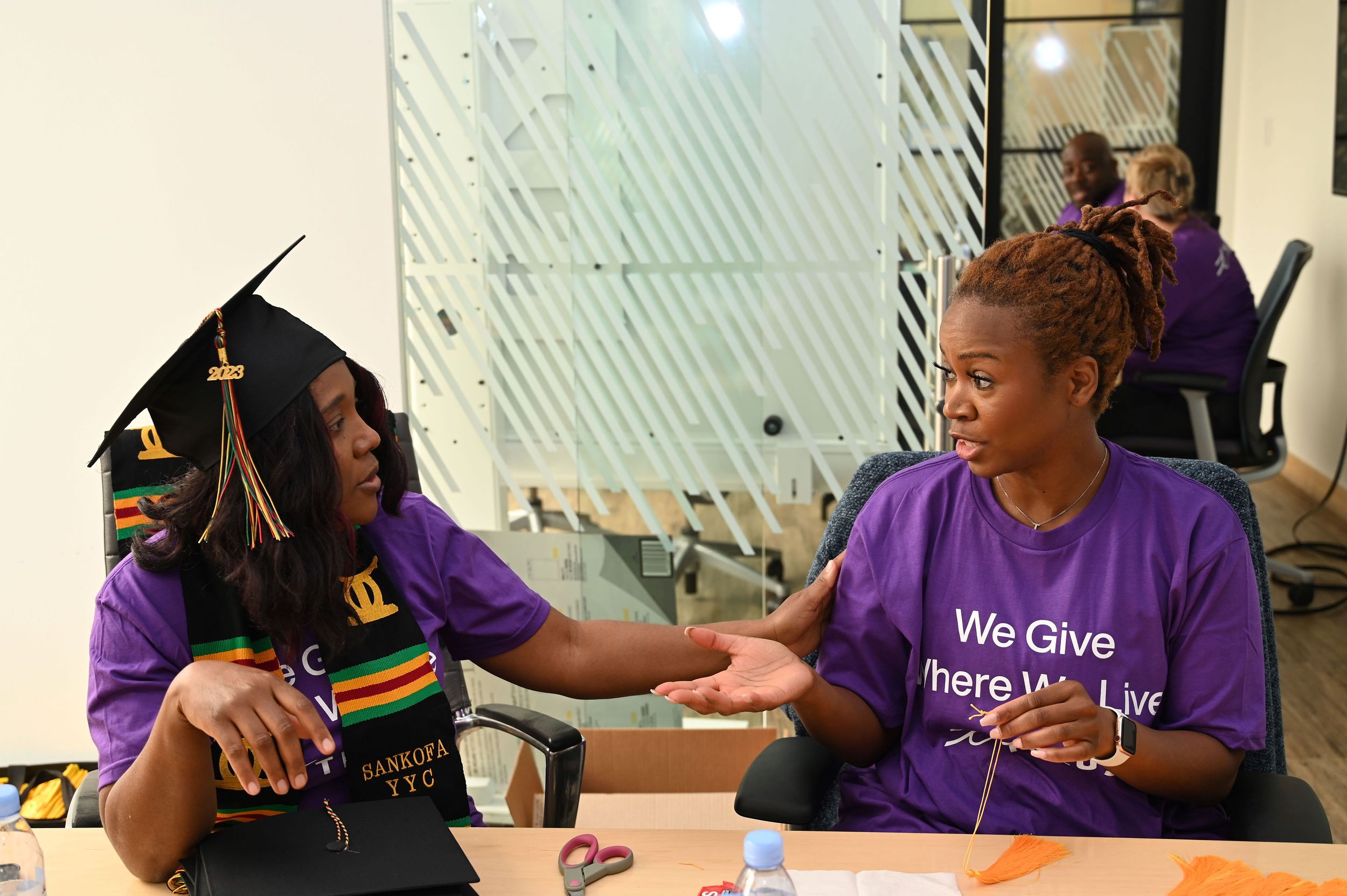 Two black women are volunteering their time on behalf of Telus. They are sitting at a table and assembling graduation caps.