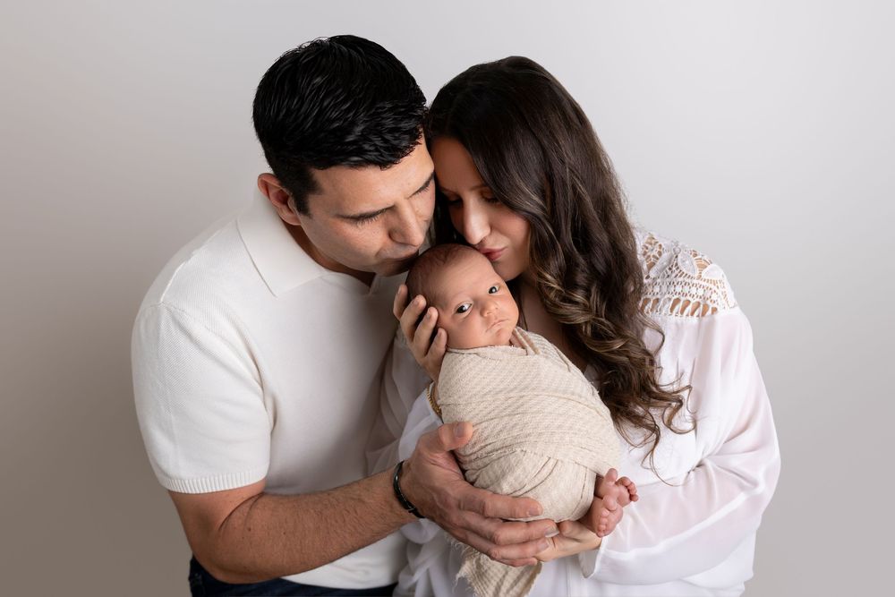 Parents tenderly embrace their sleeping newborn baby wrapped in soft beige fabric against a light gray background.