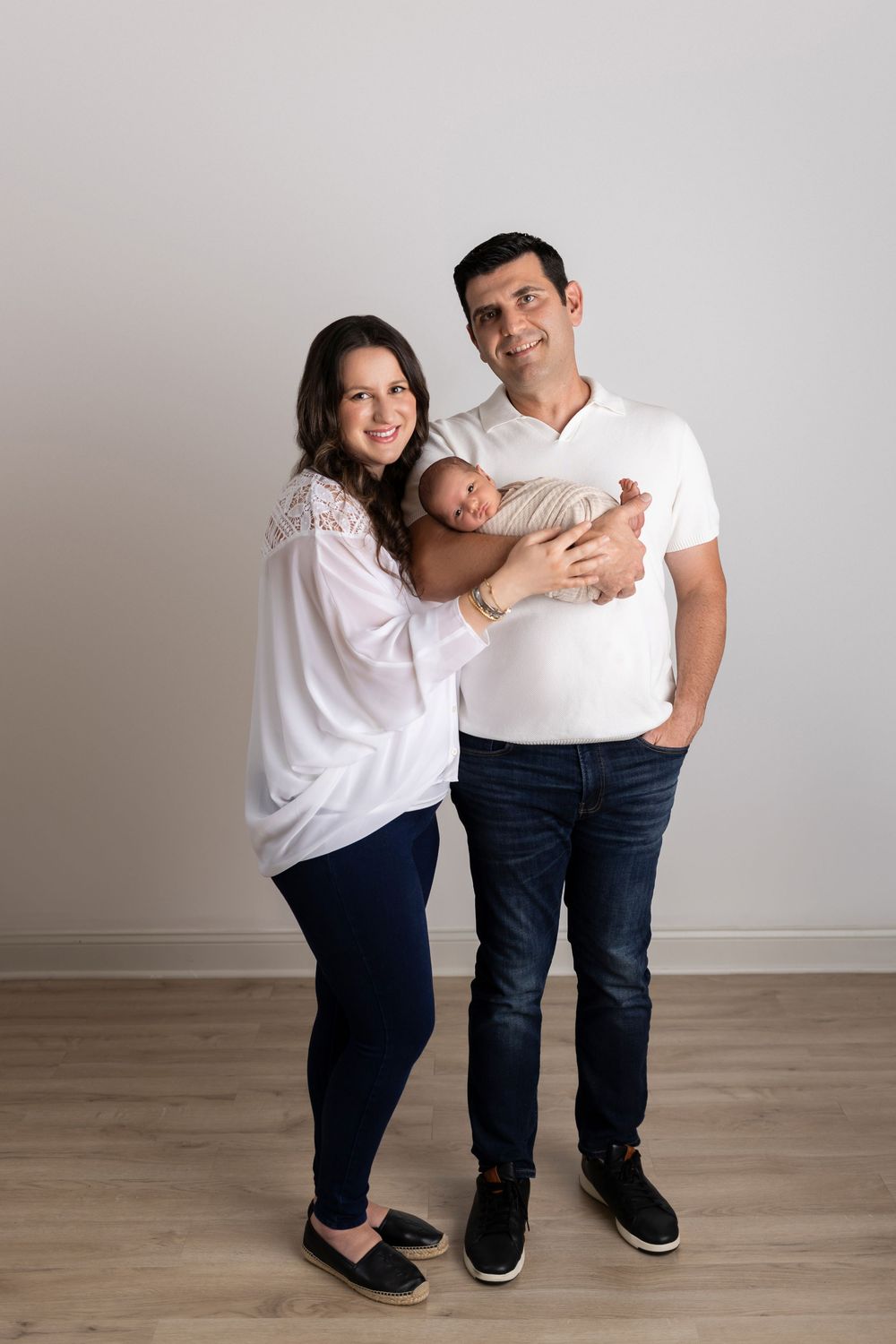 A couple wearing white tops and dark pants holds their baby while standing together in a studio setting.