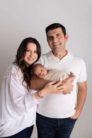 Parents in coordinated white outfits share a joyful moment with their swaddled infant against a gray backdrop.