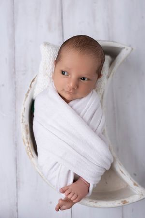 Newborn baby wrapped in white fabric rests peacefully in a curved white prop bowl.