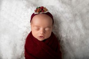 A sleeping newborn wrapped in a burgundy knit blanket on white fur wearing a delicate floral headband in New Jersey Photography Studio