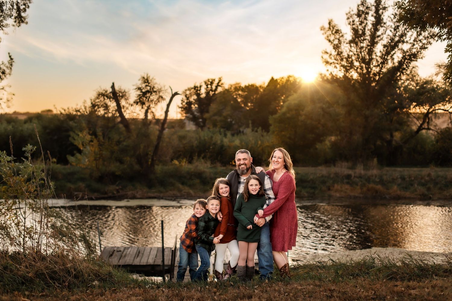 A family portrait at sunset by a reflective pond with autumn trees in the background.