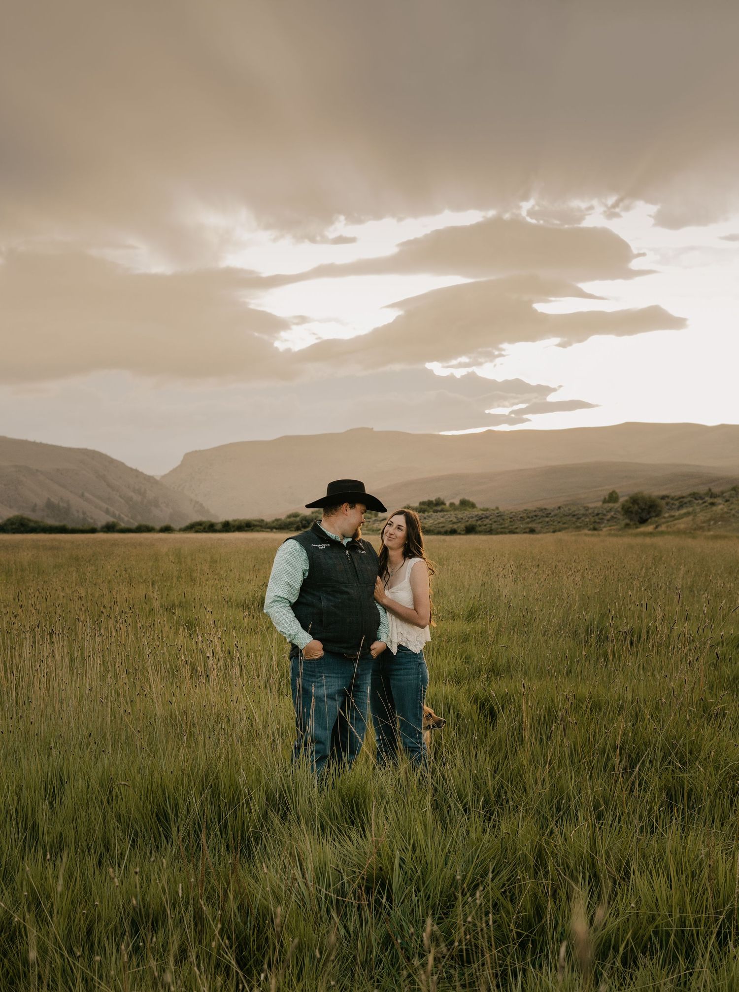 A young couple with the man on a horse leaning down the kiss the woman behind his cowboy hat. The mountains and sunset are in the background